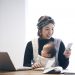 Japanese woman in casual clothes carrying her baby in baby carrier and using a smartphone on the desk. She's working and doing childcare at home.